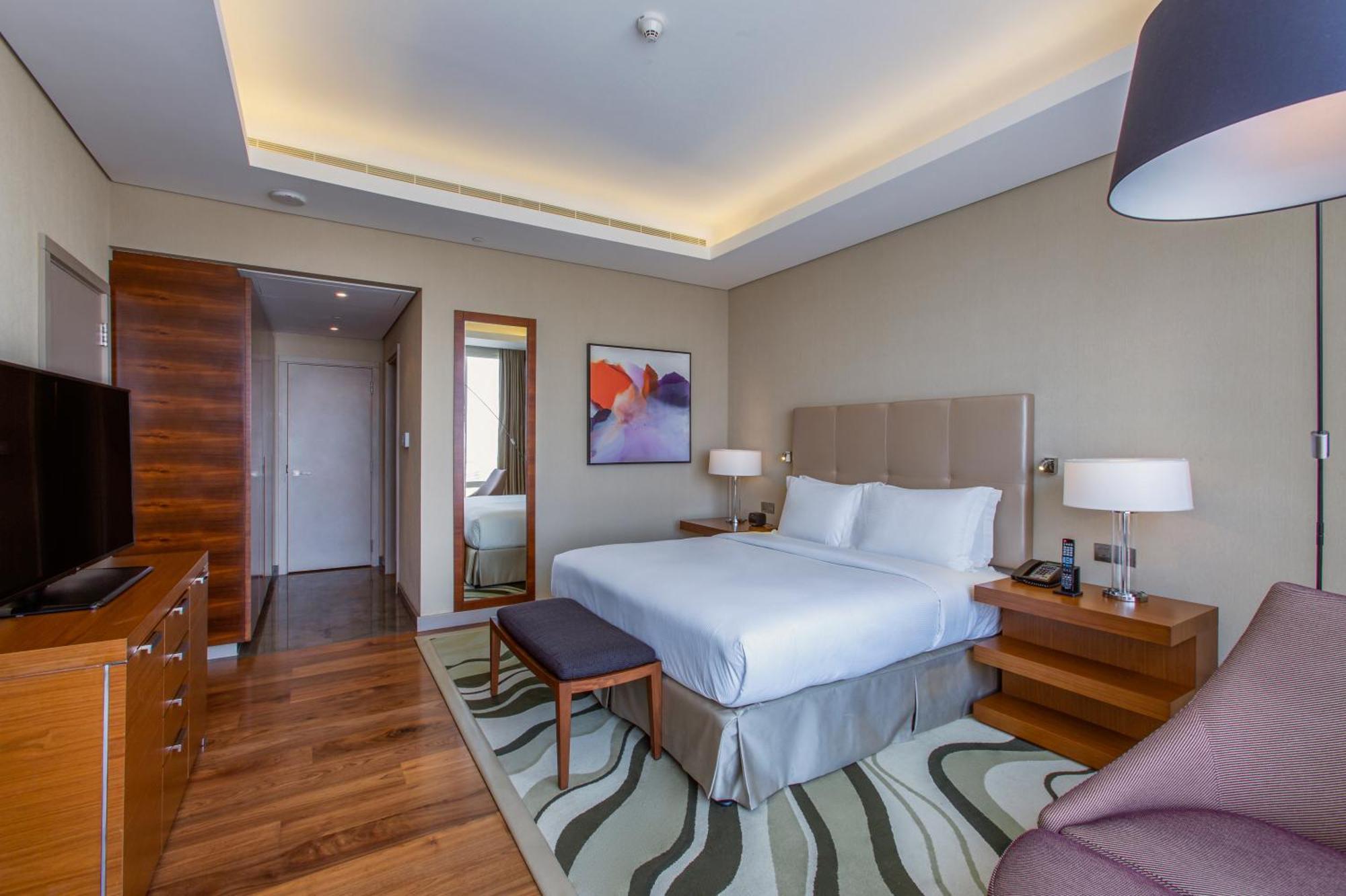 Hilton Riyadh Hotel & Residences Eksteriør bilde The photo shows a modern hotel room with a comfortable and stylish design. It features a large bed with white linens and a headboard, flanked by two bedside lamps. There is a small bench at the foot of the bed and a cozy chair in the corner. A flat-s