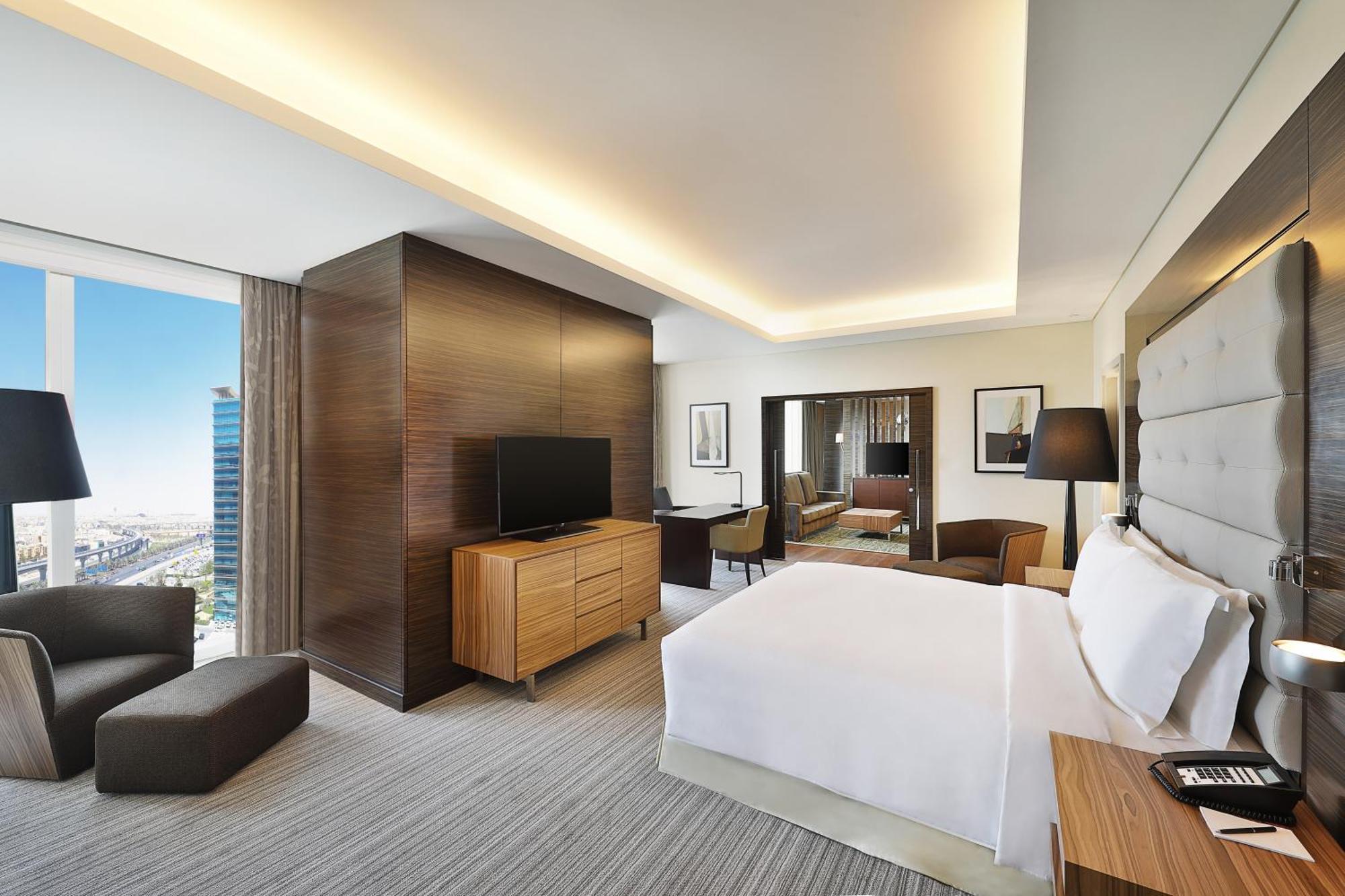 Hilton Riyadh Hotel & Residences Eksteriør bilde The photo shows a modern hotel room. It features a neatly made bed with white linens, a wooden nightstand beside it, and a cozy armchair. The room has a large window that provides a view of the outside, likely showcasing a cityscape. A flat-screen TV