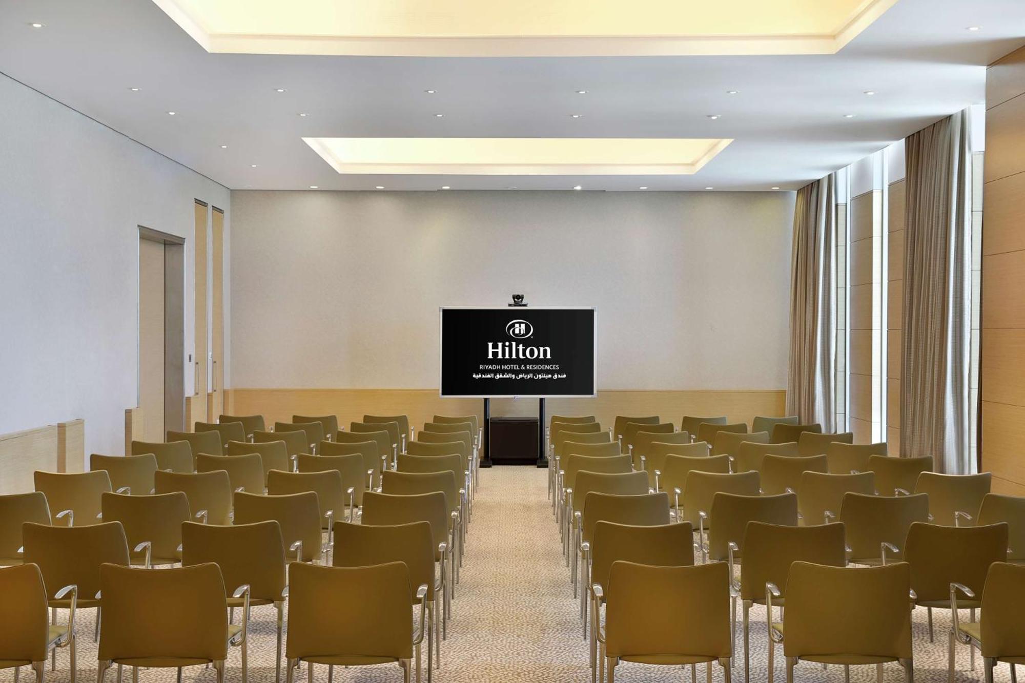 Hilton Riyadh Hotel & Residences Eksteriør bilde The photo shows a conference room or meeting space. It features rows of green chairs arranged facing a large screen that displays the Hilton logo. The room has a modern design with light-colored walls and a bright ceiling. Natural light appears to co