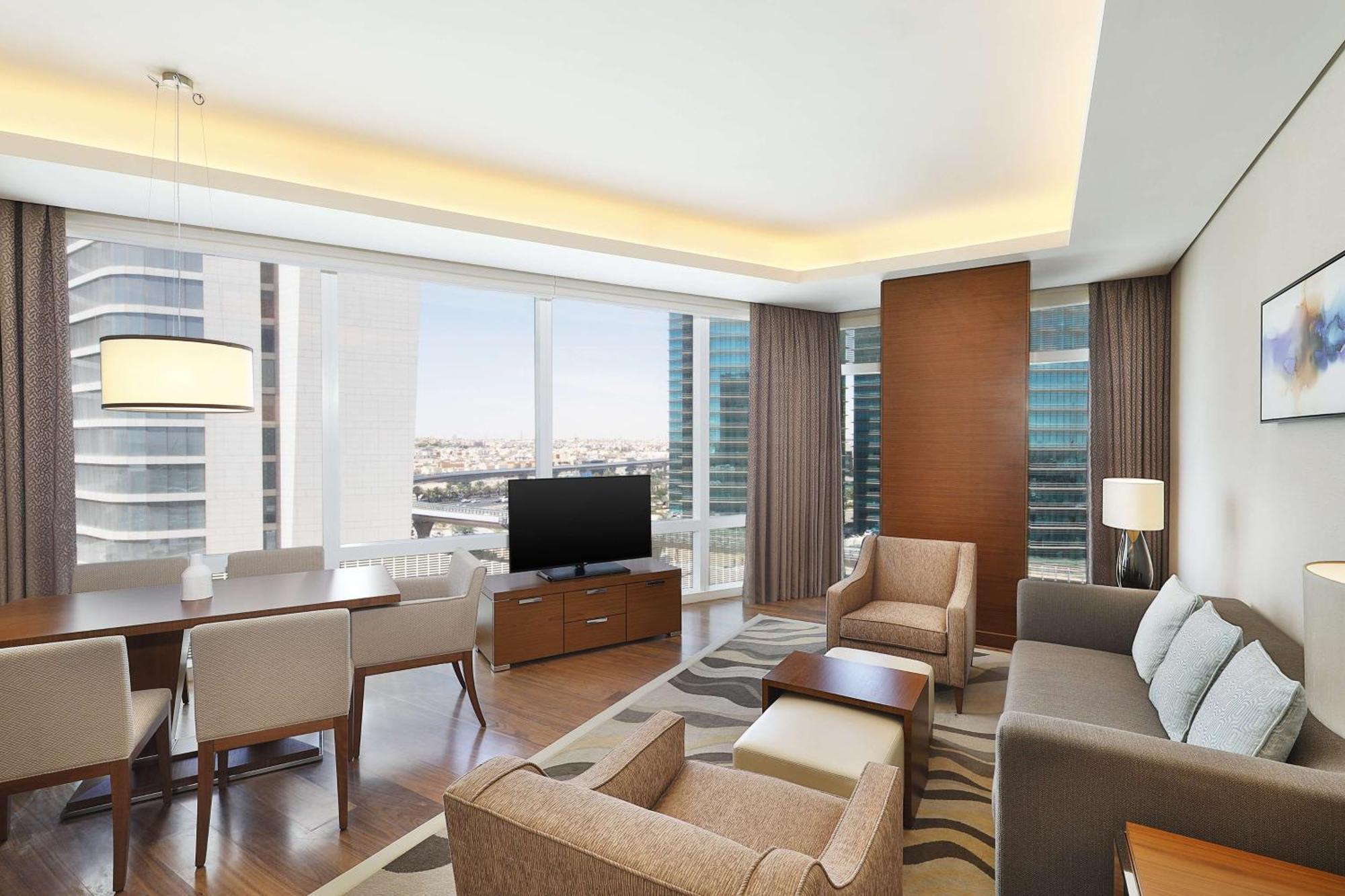 Hilton Riyadh Hotel & Residences Eksteriør bilde The image shows a modern, stylish living area in an apartment or hotel suite. It features large windows that provide a view of the cityscape, allowing natural light to flood the space. The furnishings include a comfortable sofa, armchairs, and a coff