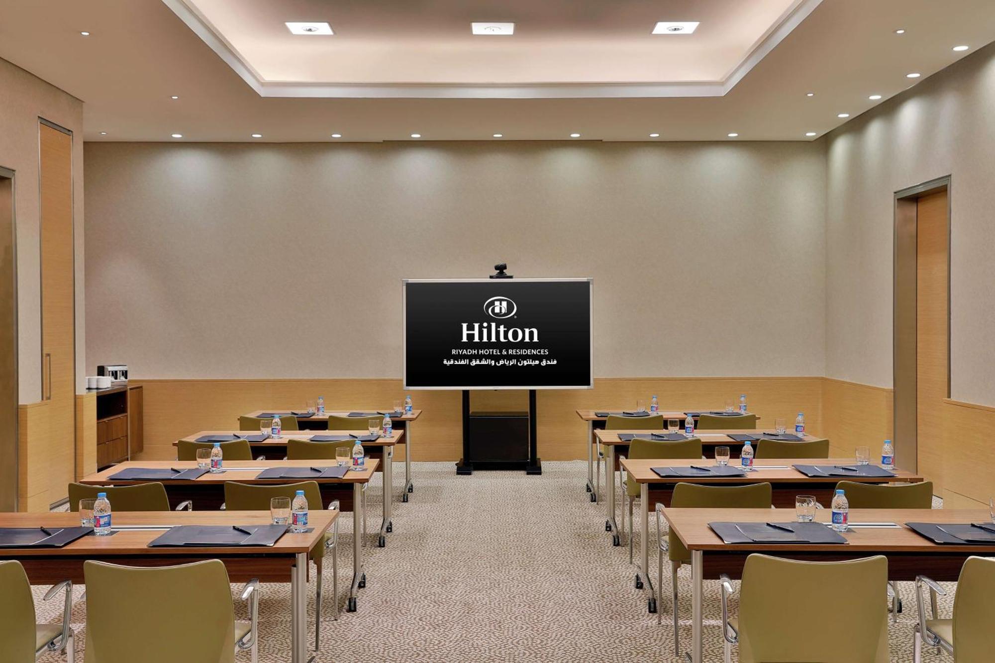Hilton Riyadh Hotel & Residences Eksteriør bilde The photo shows a conference or meeting room set up for a presentation. There are several tables arranged in a classroom style, each equipped with chairs. On each table, there are notepads and water bottles. At the front of the room is a large screen