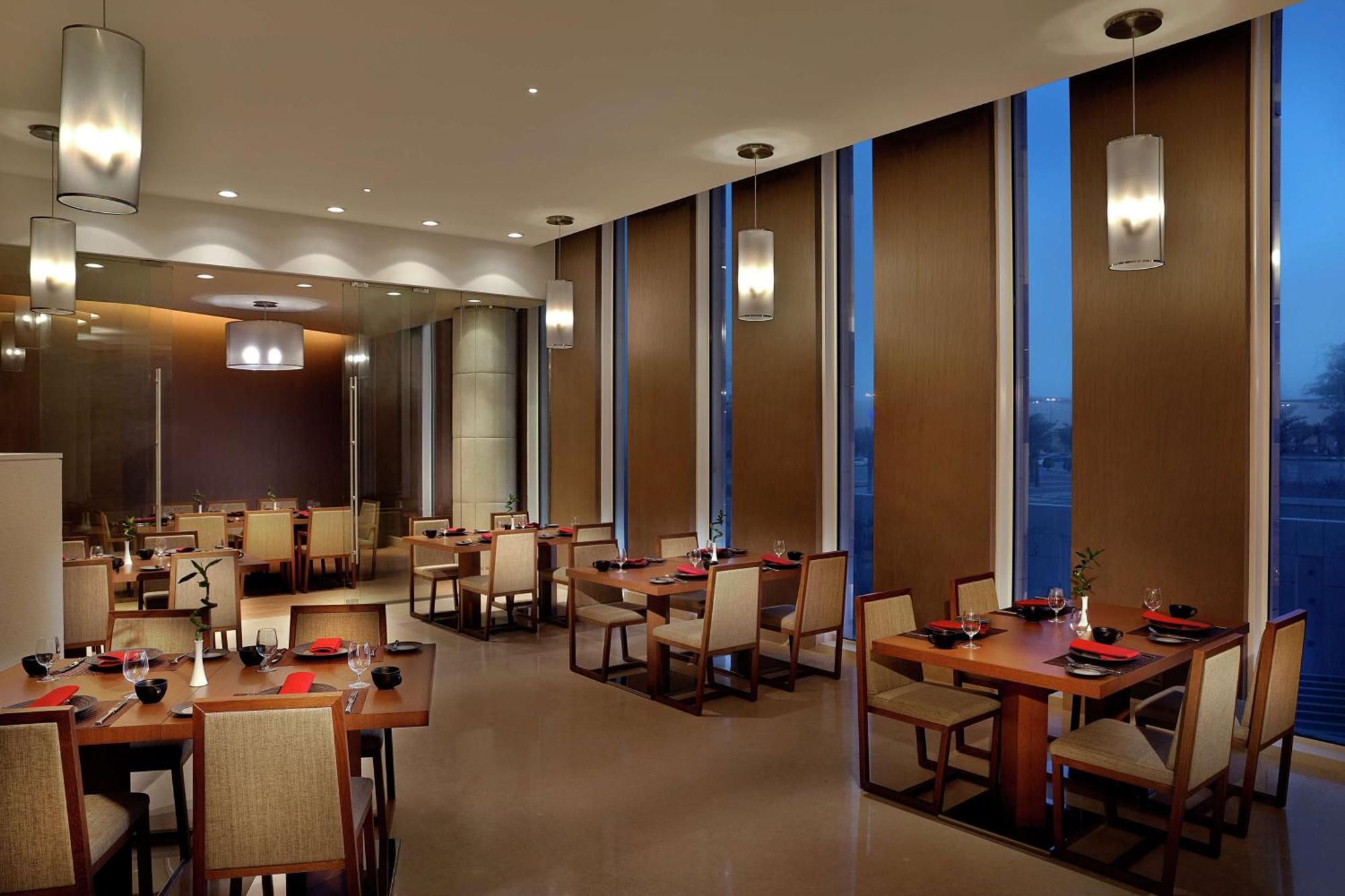 Hilton Riyadh Hotel & Residences Eksteriør bilde The image depicts a modern restaurant interior. It features several dining tables arranged neatly, each set with plates and utensils. The room has large windows with blinds partially drawn, allowing some ambient light to filter in, while evening ligh