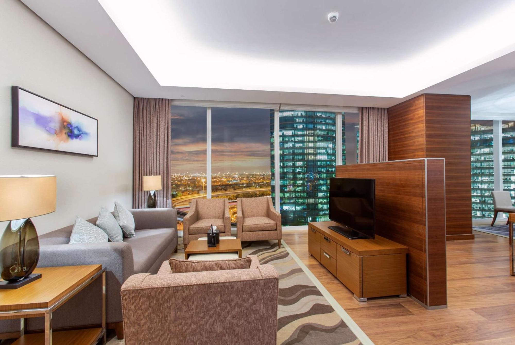 Hilton Riyadh Hotel & Residences Eksteriør bilde The photo shows a modern living room in a stylish apartment. The space features a comfortable seating arrangement with a light gray sofa and two brown armchairs, complemented by a wooden coffee table. There is a large flat-screen TV mounted on a wood