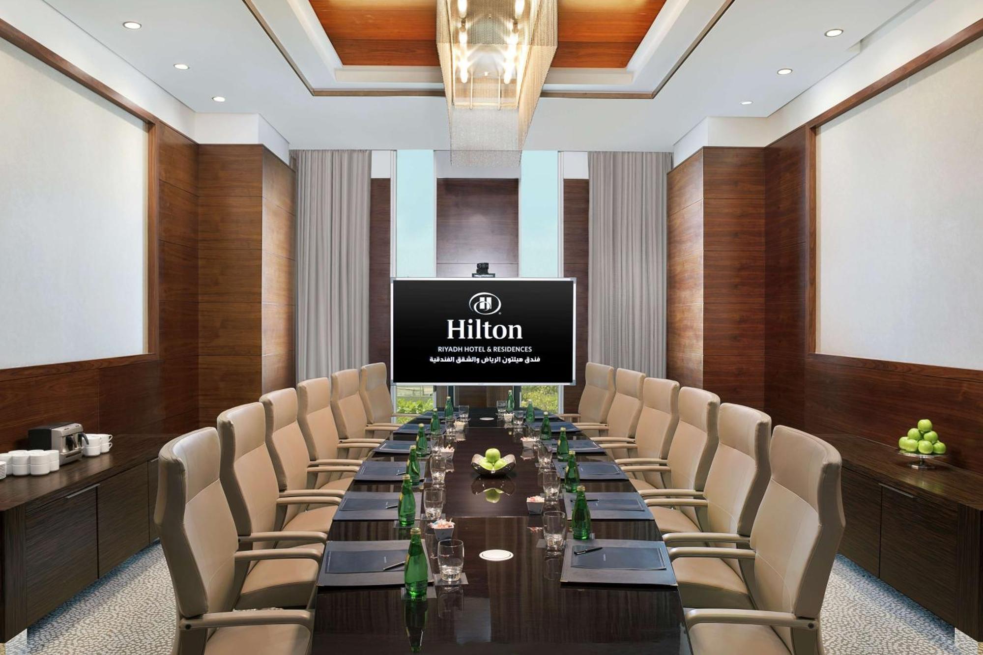 Hilton Riyadh Hotel & Residences Eksteriør bilde The photo shows a modern conference room within a Hilton hotel. There is a large wooden table surrounded by several comfortable chairs. At the head of the table is a large screen displaying the Hilton logo. The room is well-lit with stylish overhead 
