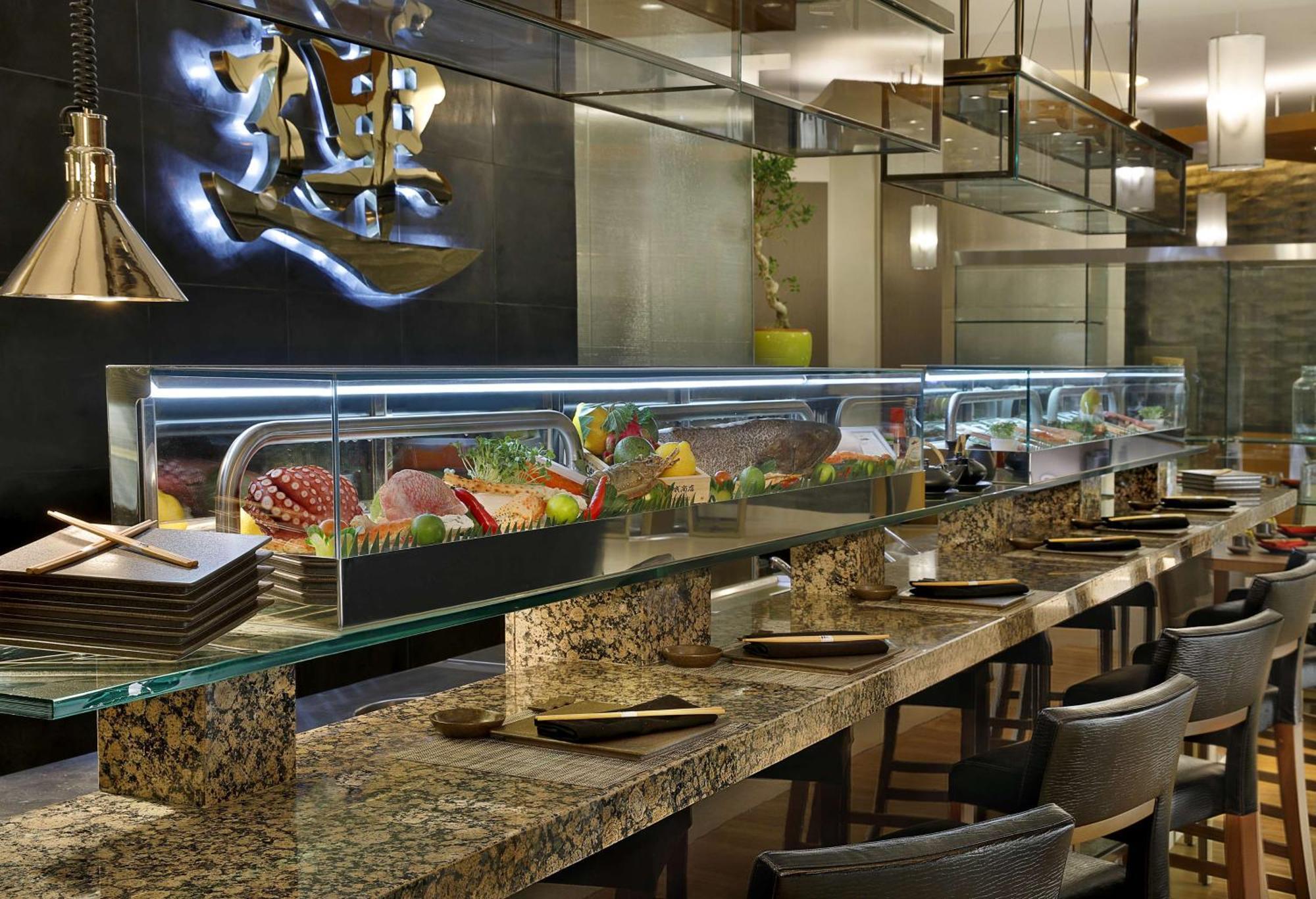 Hilton Riyadh Hotel & Residences Eksteriør bilde The photo shows an interior view of a sushi restaurant. There is a long, sleek sushi bar with a glass display case filled with various sushi and sashimi offerings. The counter is made of polished stone, and there are several dark chairs arranged neat